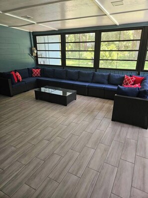A modern living room with a sectional sofa, a glass coffee table, and large windows offering a view of greenery. - Private Pool • Sleeps 16 • Huge Yard • AC GameRoom (Lakeland)