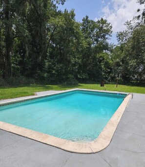 Piscina retangular com borda revestida, rodeada por um deck de concreto e uma área gramada com árvores.