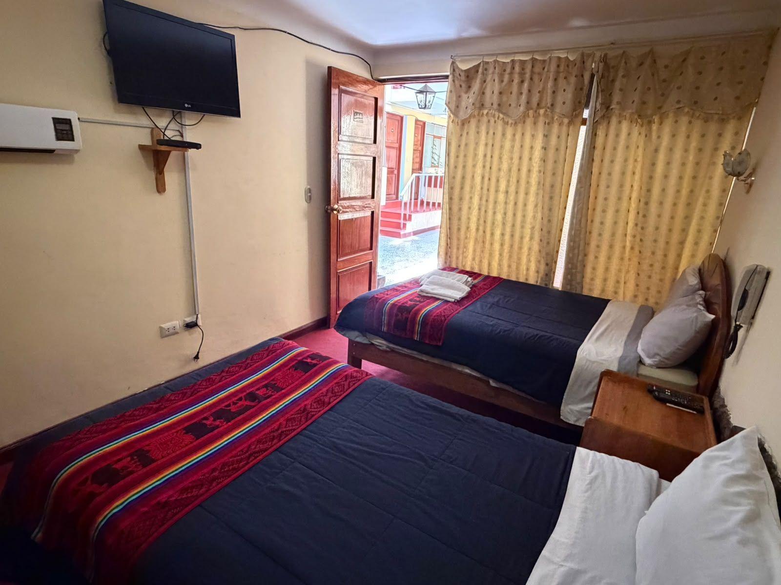 A room with two beds, a television mounted on the wall, and a window with curtains.