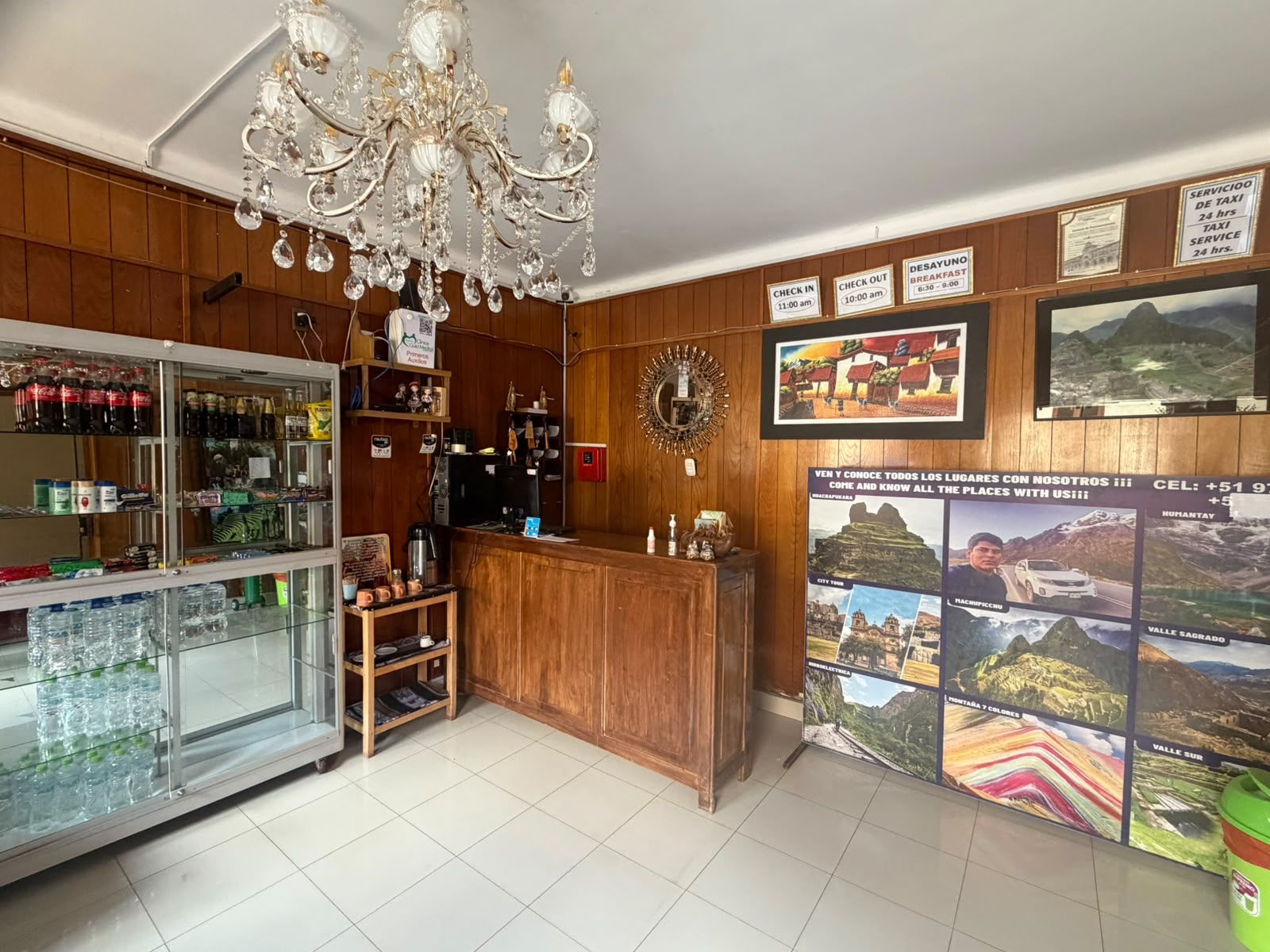 A hotel lobby with a chandelier, a reception desk, a refrigerator stocked with beverages, and a wall with informational posters.