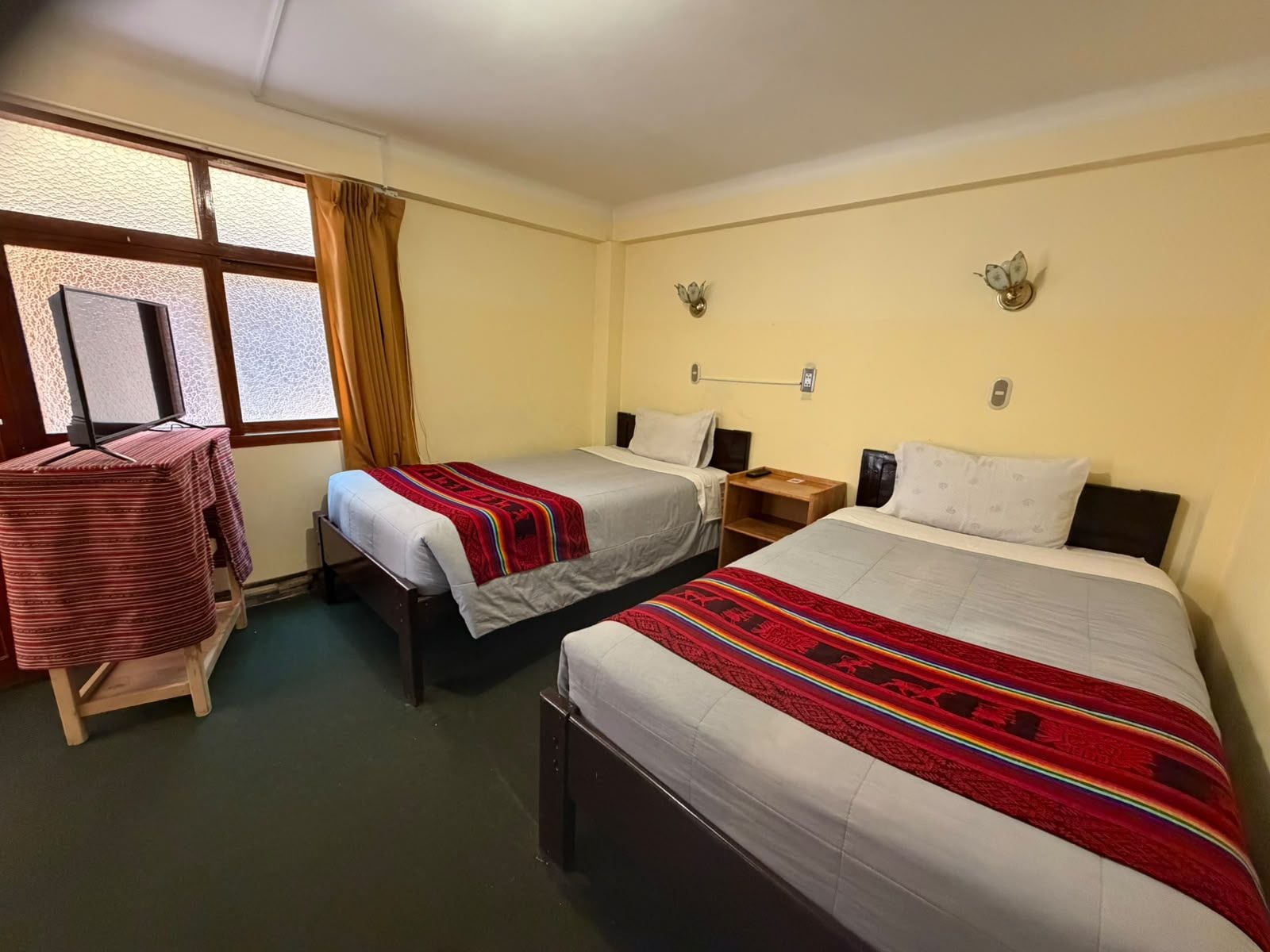 Una camera d'albergo con due letti singoli, un tavolino e una televisione, e una finestra con tende.