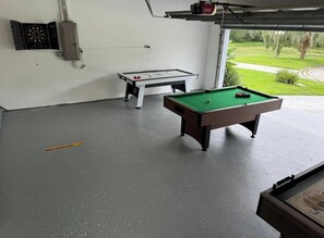 A billiards table, a dartboard, and a foosball table in a room with a concrete floor and a view of a green landscape through a garage door. - 3 Bedroom Spacious AC Pool Home w/ Gameroom (Lakeland)