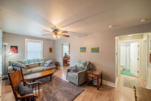Ein Wohnzimmer mit Sofa, Sessel, Couchtisch und Deckenventilator. Im Hintergrund ist ein Essbereich mit Tisch und Stühlen zu sehen.