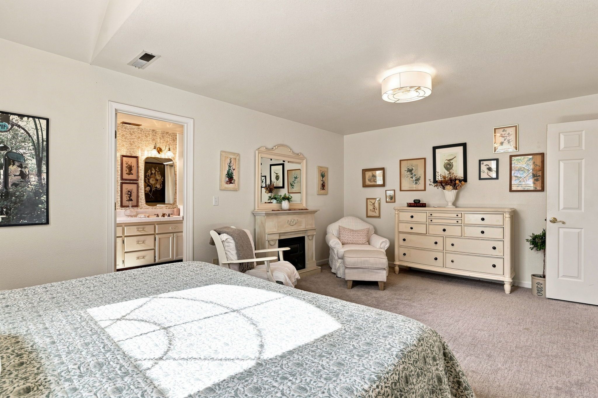 A bedroom with a bed, dresser, chair, fireplace, and wall-mounted artwork.