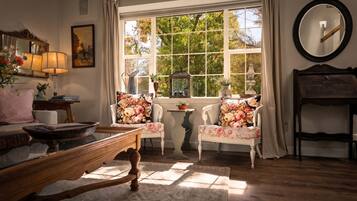 A cozy room with a large window, a wooden table, two chairs with floral upholstery, a mirror, and a lamp.