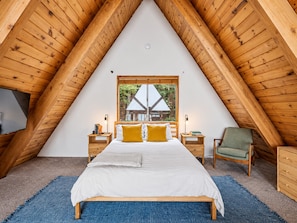 Ett sovrum med ett sluttande tak, en säng med vita sängkläder, utsikt över ett hus genom ett fönster och möbler i trä.