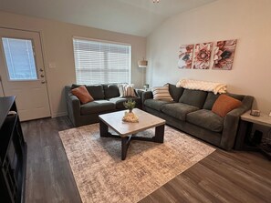 A living room with a sofa, a coffee table, and a rug. There is a door and a window with blinds. - Sage and Stillness Extended Stay Retreat (Killeen)