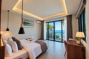 A hotel room with a bed, bedside lamps, a desk, a chair, and a view of the sea. - 3-Bedroom Beach Front Villa with Ocean View in Tulamben Karangasem Bali (Karangasem)