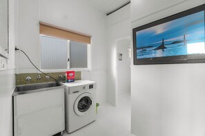 Un espace buanderie compact comprenant un lave-linge, un évier et une fenêtre avec des stores.