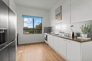 A modern kitchen with a wooden floor, white cabinets, a stainless steel refrigerator, a built-in oven, and a window with a view of greenery. - Beach Shack Escape Beach Just Across the Road (Aldinga Beach)