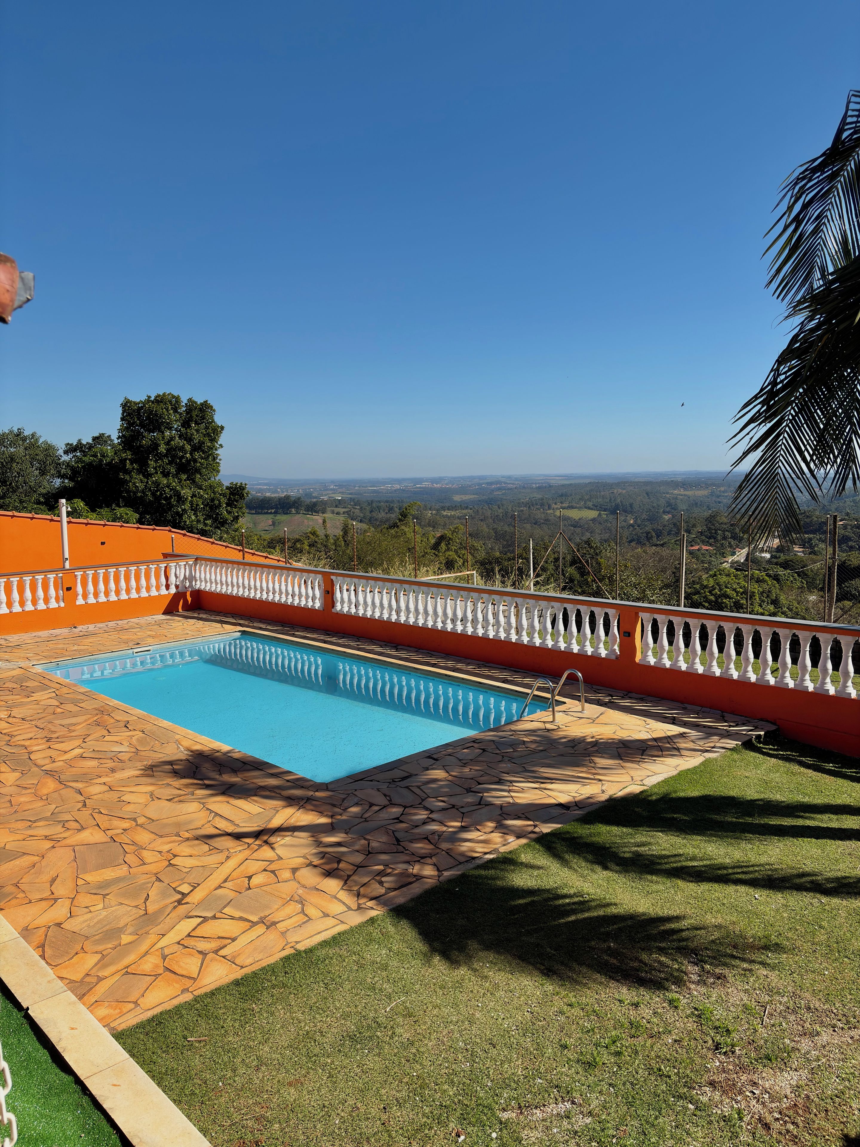 A swimming pool with a stone-tiled deck and a white balustrade, surrounded by grass and trees, with a view of distant hills.