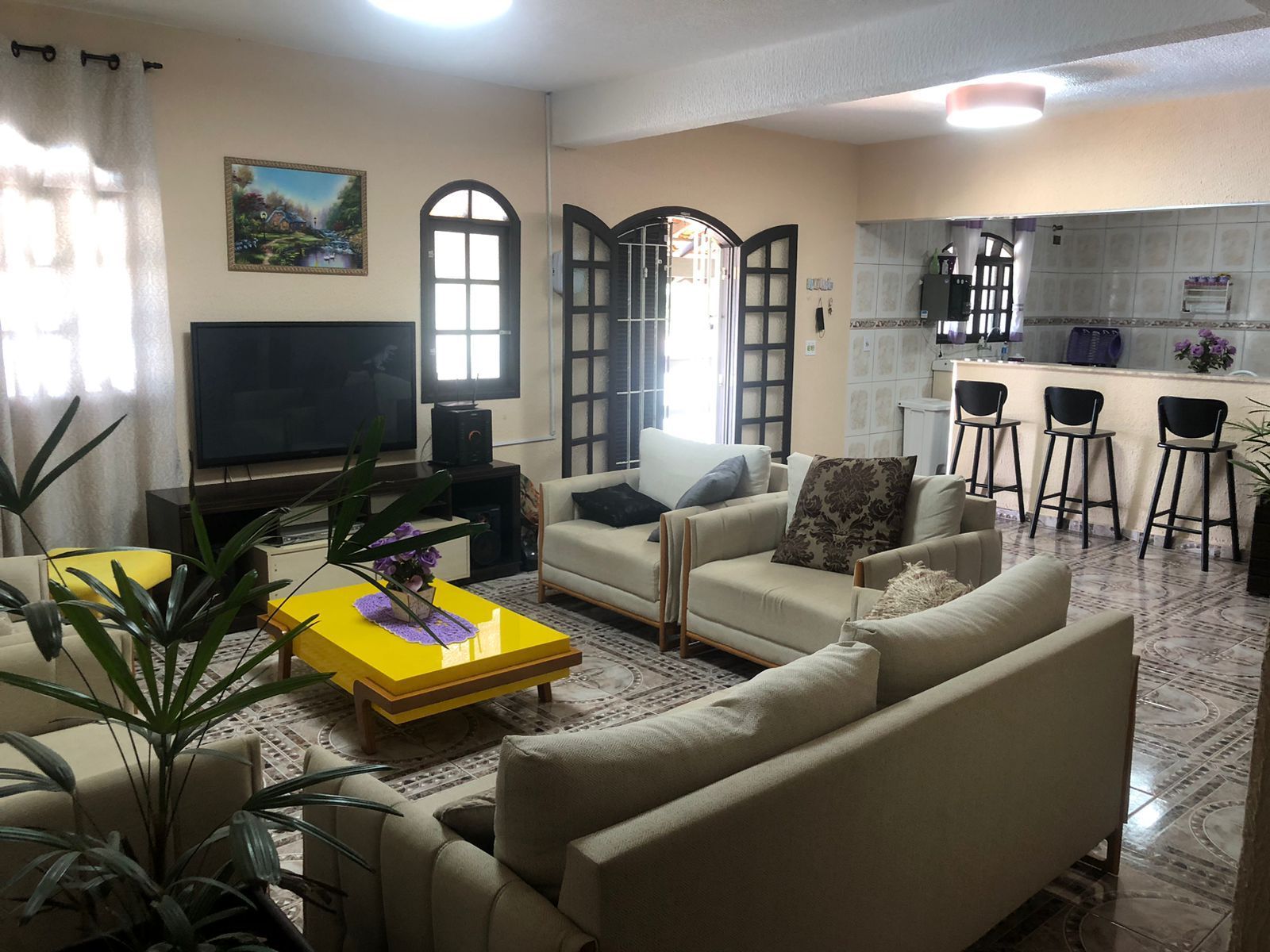 A living room with a sofa, a coffee table, and a TV. There is a kitchen area with a bar and stools.