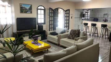 A living room with a sofa, a coffee table, and a TV. There is a kitchen area with a bar and stools.