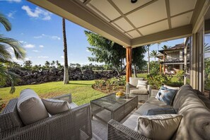 Een overdekt terras met rieten meubilair en een glazen bijzettafeltje, met uitzicht op een aangelegde tuin met palmboompjes en een stenen muur.