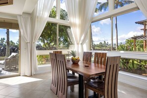 A dining area with a wooden table and chairs, large windows with curtains, and a view of tropical scenery. - NEW Mauna Lani Villa Sunset Designer Renovated (Waimea)