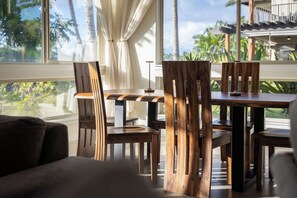 Een eetruimte met een houten tafel en stoelen, grote ramen met gordijnen, en uitzicht op palmbomen.