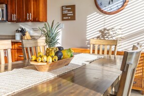 A wooden dining table with a fruit bowl in the center, a clock on the wall, and a kitchen area in the background. - Pool & Ocean View Villa: 3BD | Sunset Kona Side (Kailua-Kona)