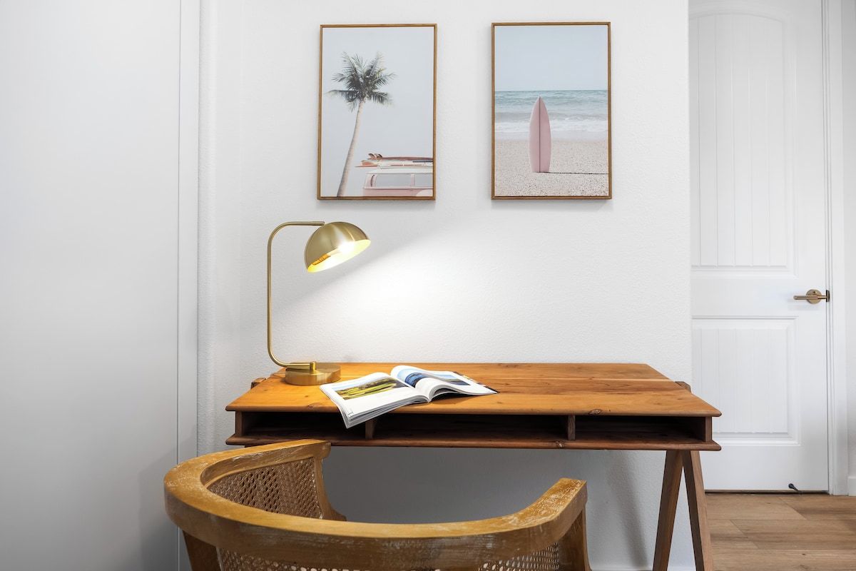 A room with a wooden desk, a wicker chair, a lamp, and two framed pictures on the wall.