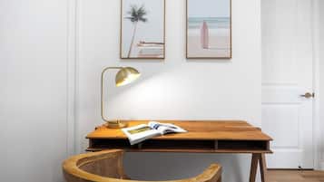 A room with a wooden desk, a wicker chair, a lamp, and two framed pictures on the wall.