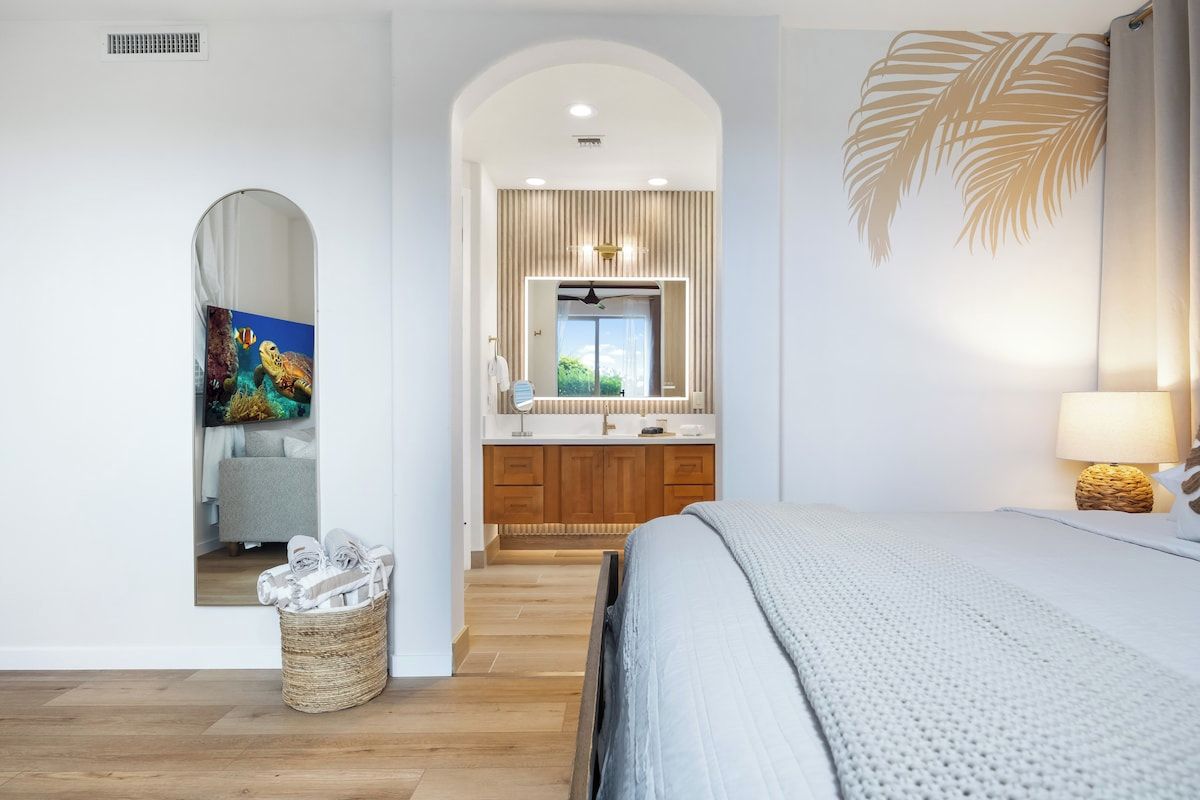 A bedroom with a large bed, a wooden floor, a mirror above the sink, and a view of the bathroom.