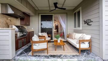 A covered patio with a grill, two white sofas, a wooden coffee table, and a ceiling fan.