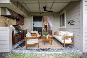 Eine überdachte Terrasse mit Grill, zwei weißen Sofas, einem Holztisch und einem Deckenventilator.