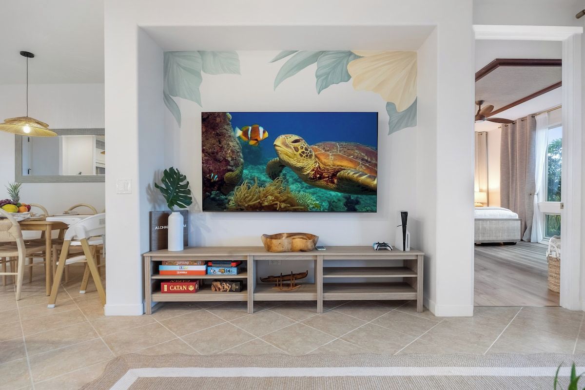 A living room with a large flat-screen TV mounted on a wall, a wooden entertainment unit below, and a dining area with a table and chairs.