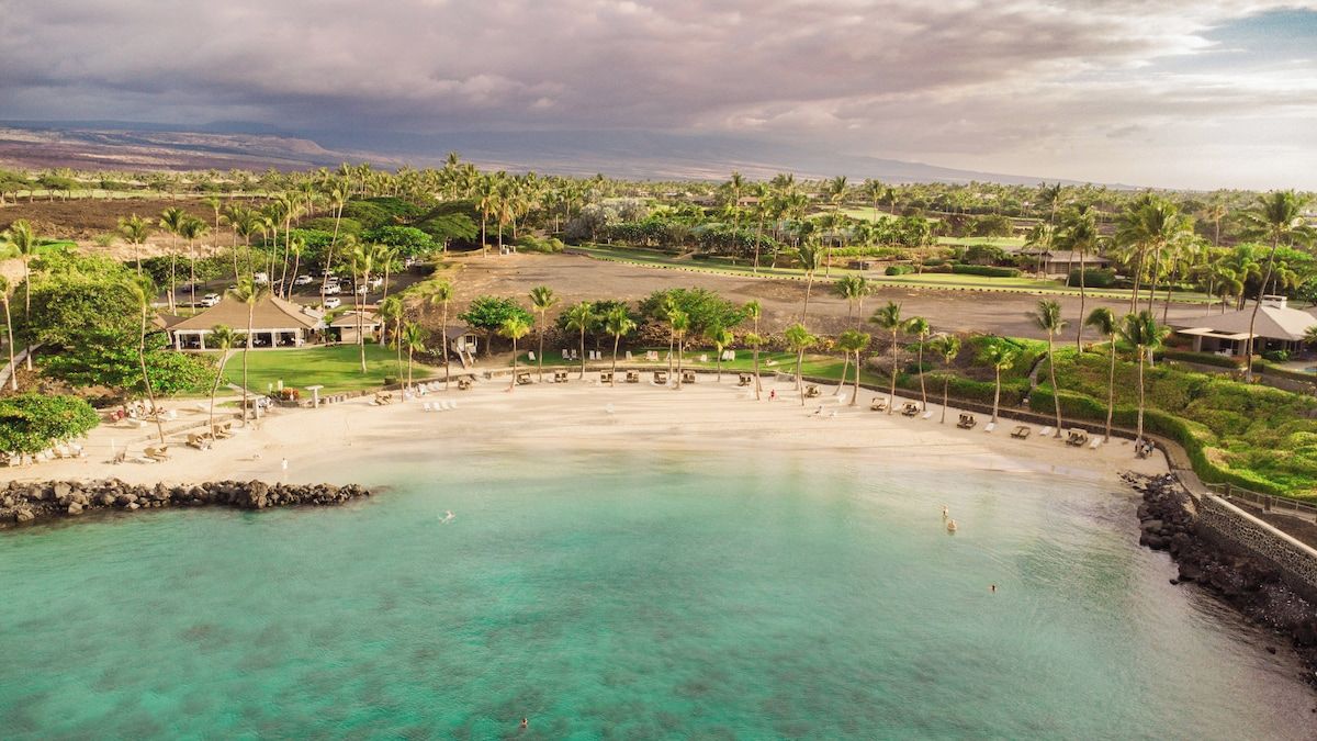 A coastal resort with a clear turquoise beach, palm trees, and a golf course in the background.