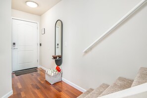 A hallway with a white door, a wall-mounted mirror, a small shelf with bowls, and a basket with items. - Upgraded 3 bed w Ocean View, Pool, Hot tub, 2 King (Waikoloa Village)