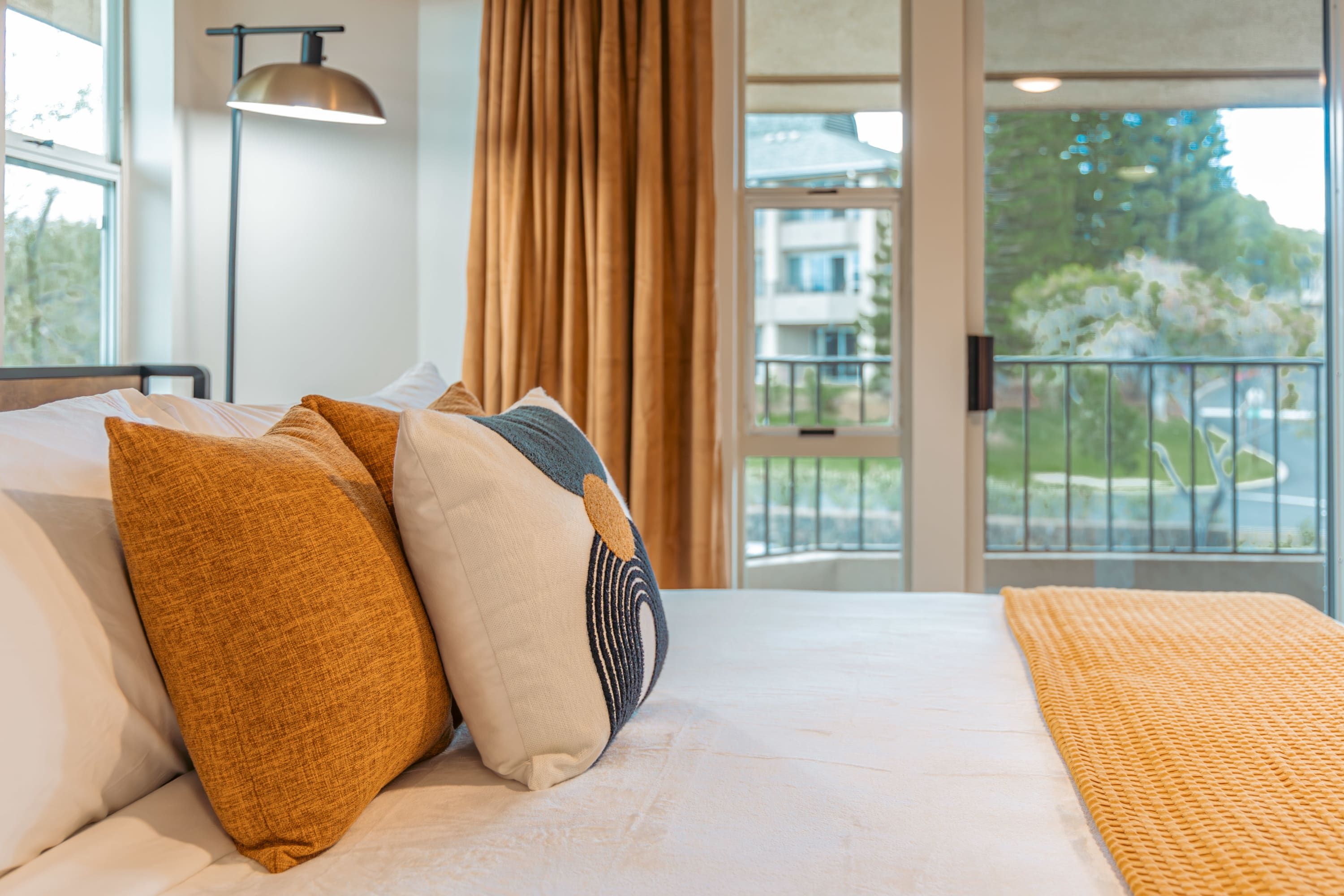 A hotel room with a bed, pillows, a lamp, curtains, and a view of the outside.