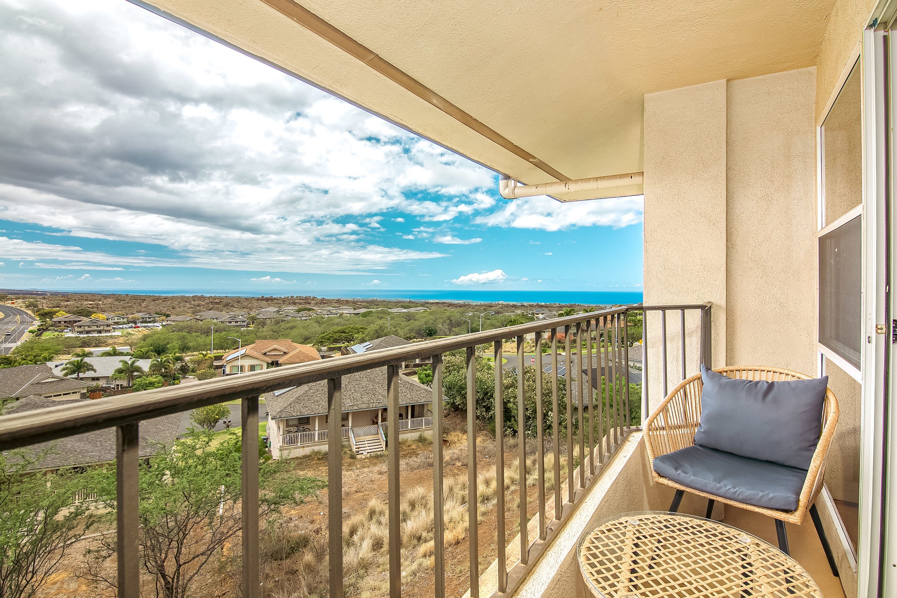 Un balcón con una silla de mimbre y un cojín, con vista a una zona residencial con casas y cielo despejado.