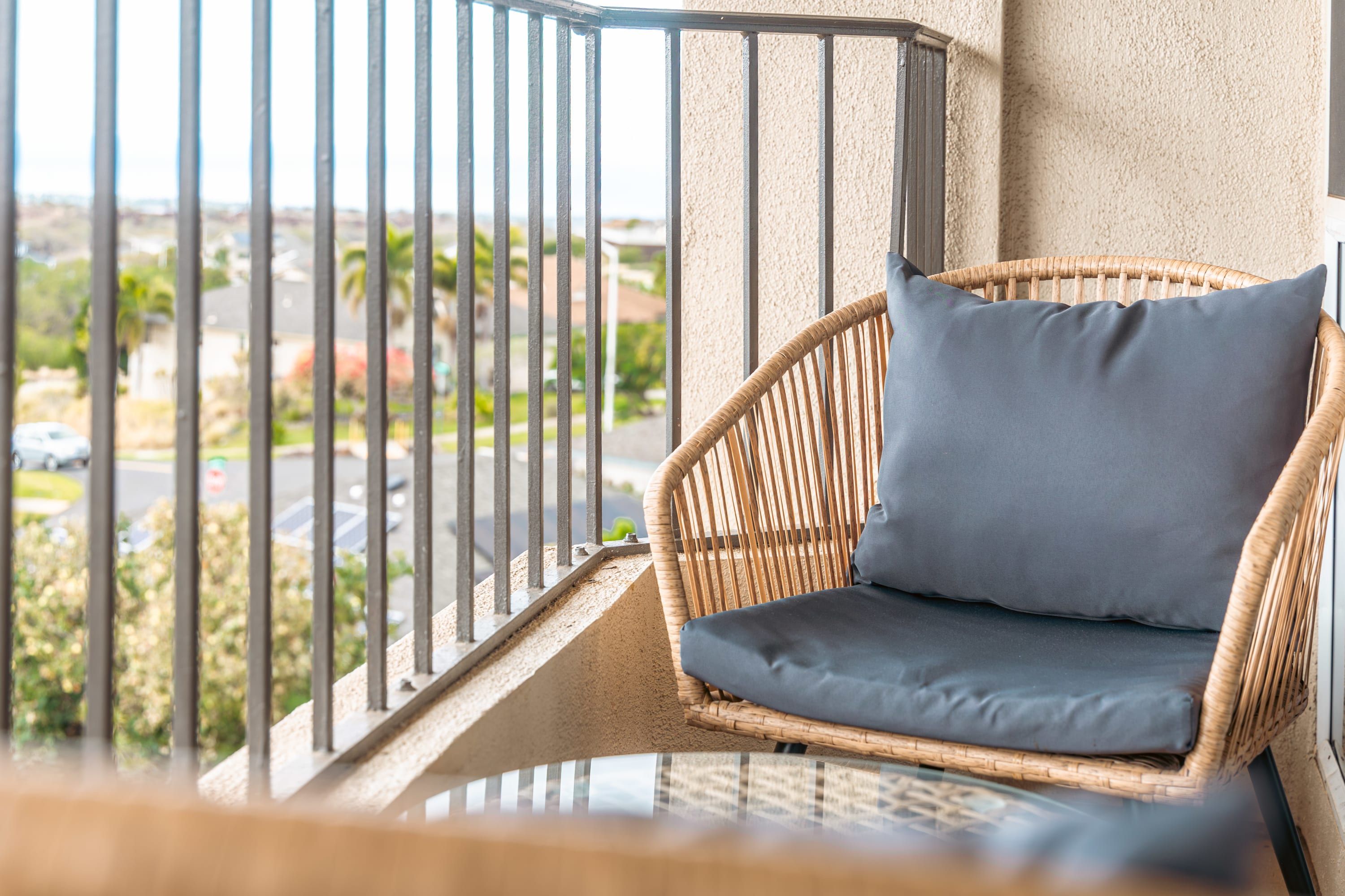 A wicker chair with a cushion on a balcony with a metal railing, overlooking a street with cars and greenery.