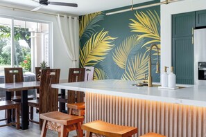 A modern kitchen with a central island, wooden chairs, and a view of the outdoors. - Treasure Retreat 3 bed Villa, Kings, Stylish Remodel (Waikoloa Village)