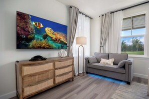 A living room with a large flat-screen TV, a grey sofa, a wooden console table, and a floor lamp. - Treasure Retreat 3 bed Villa, Kings, Stylish Remodel (Waikoloa Village)