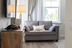 A neutral-toned sofa with a cushion, a wooden side table with a lamp, and a window with curtains. - Treasure Retreat 3 bed Villa, Kings, Stylish Remodel (Waikoloa Village)