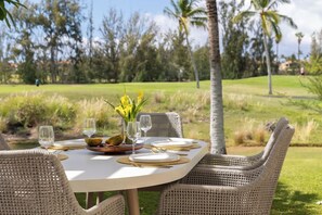 Mesa de refeições ao ar livre com pratos brancos, talheres dourados e copos, rodeada por cadeiras de vime, com vista para um campo de golfe e palmeiras ao fundo.