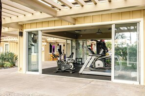 Ein überdachter Outdoor-Fitnessbereich mit Trainingsgeräten, darunter Laufbänder und stationäre Fahrräder, und einem Glastür-Eingang.