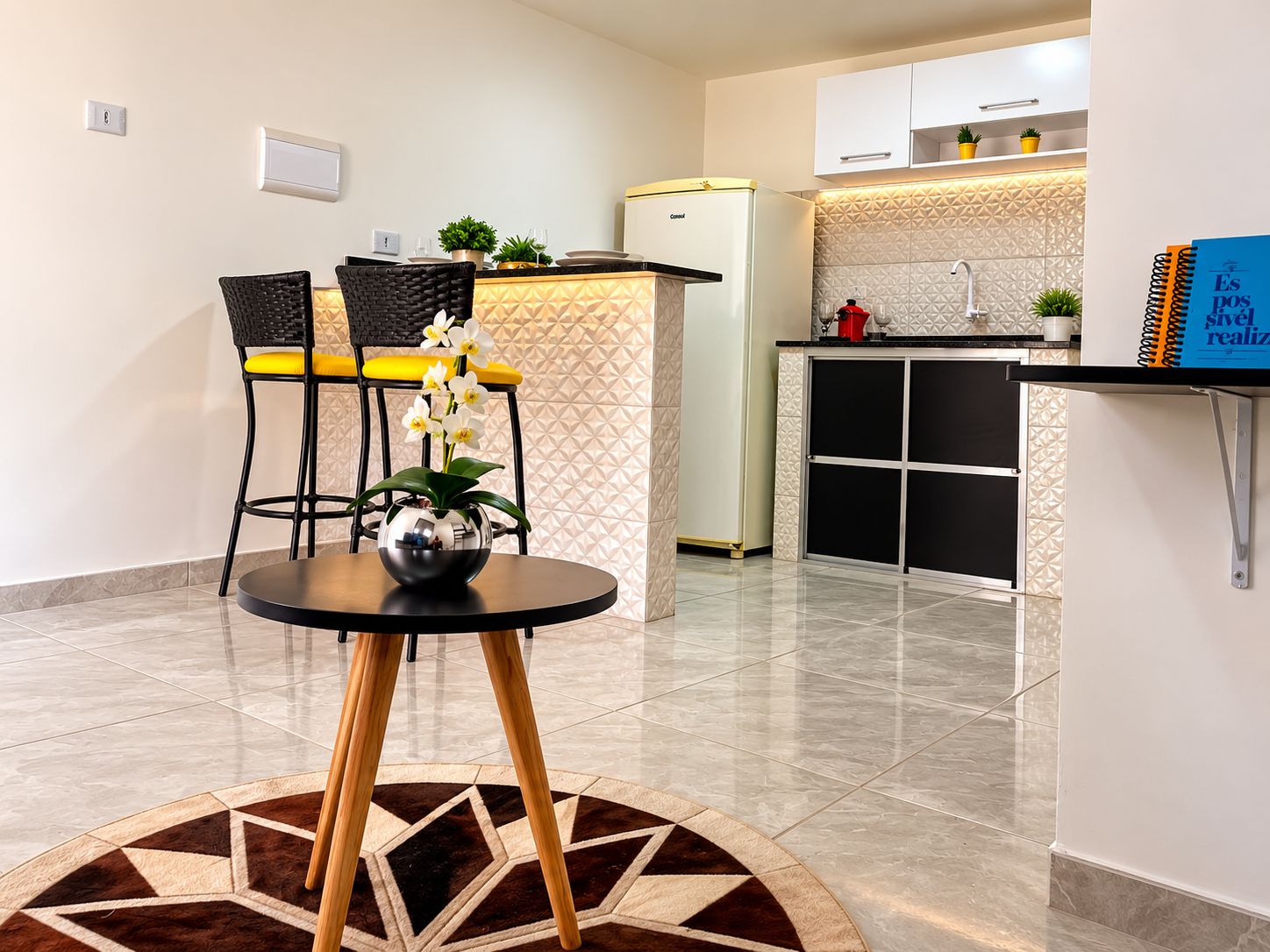 Una cocina compacta con refrigerador, un lavamanos y una encimera. Hay un comedor con mesa y sillas, y un jarrón decorativo con flores.