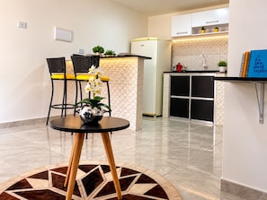 Una cocina compacta con refrigerador, un lavamanos y una encimera. Hay un comedor con mesa y sillas, y un jarrón decorativo con flores.