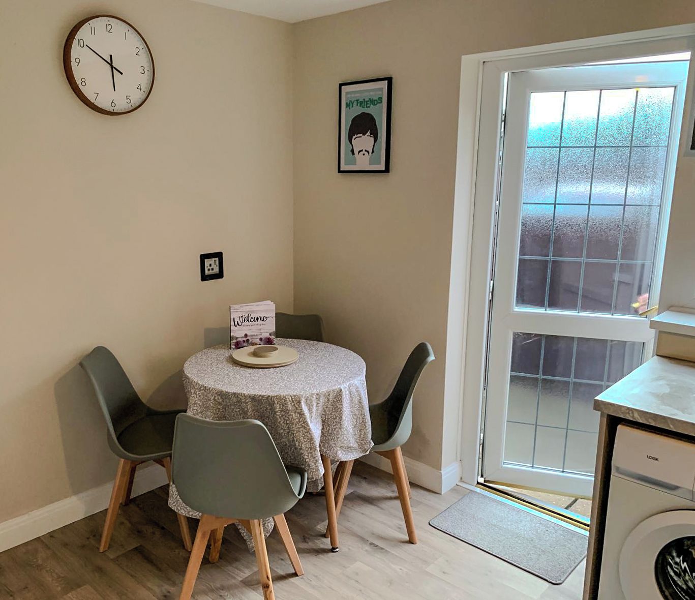A dining area with a round table, four chairs, a clock, a framed picture, and a washing machine.