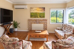 A living room with a flat-screen TV, a brick fireplace, floral sofas, and a large window with a view of greenery. - Ferguson House – Cosy Hunter Valley Escape (Cessnock)