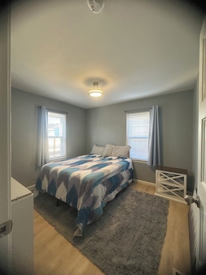Un dormitorio con una cama, una mesita de noche pequeña, un radiador y una ventana con cortinas.