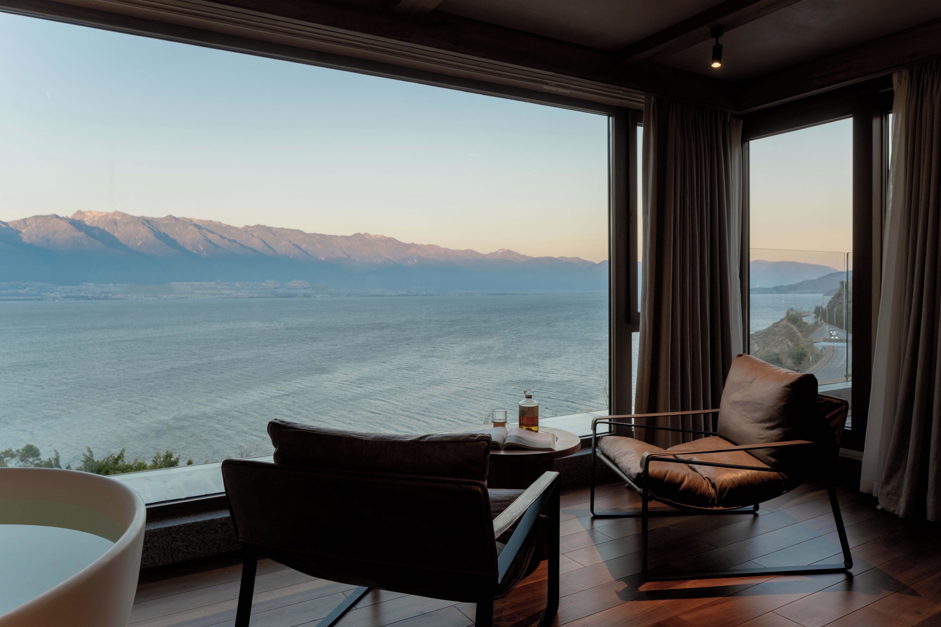 A room with a large window overlooking a body of water and mountains. There is a chair, a table with a book and a bottle, and a bathtub.