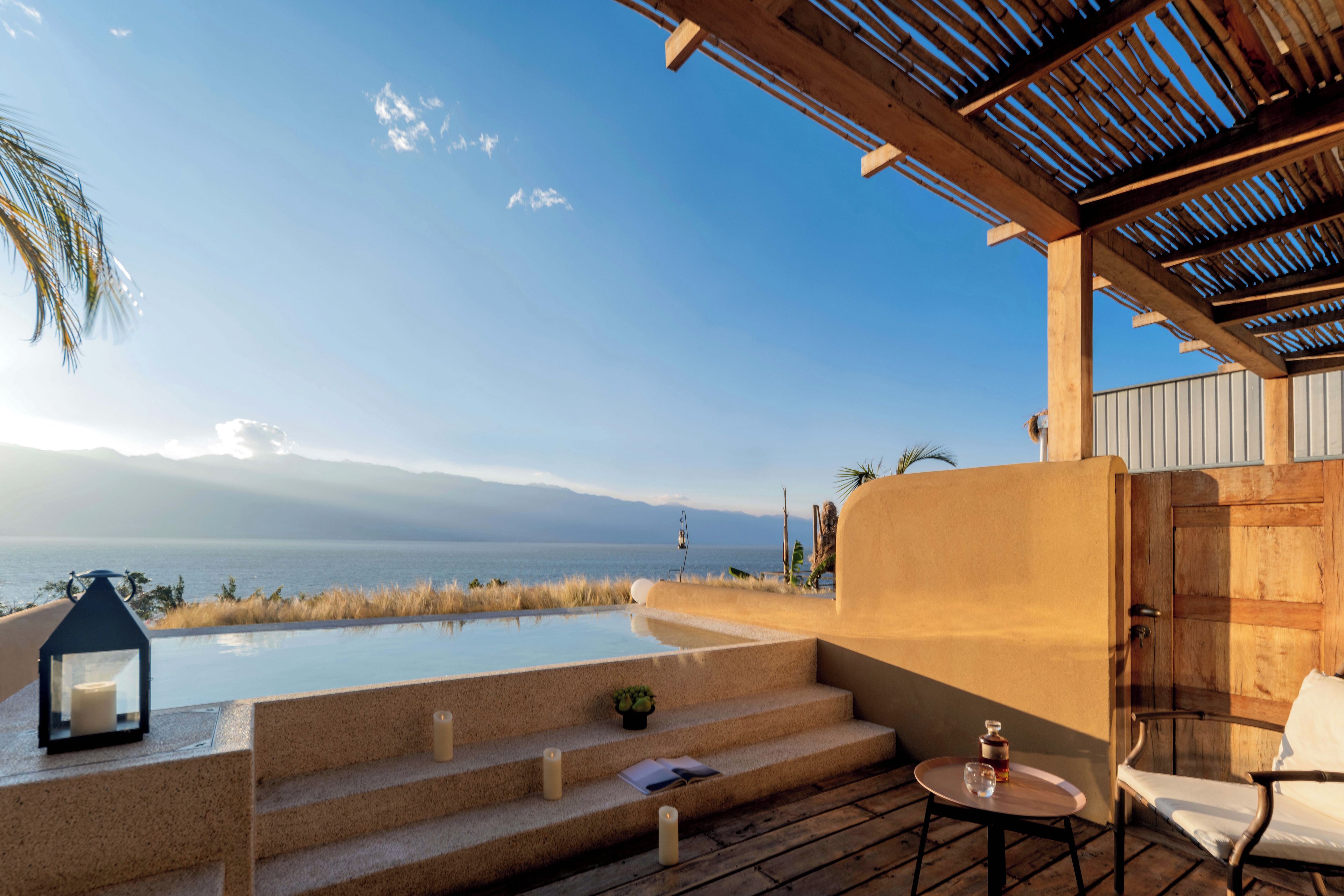 A pool area with steps leading to a lounge, overlooking a body of water and mountains.
