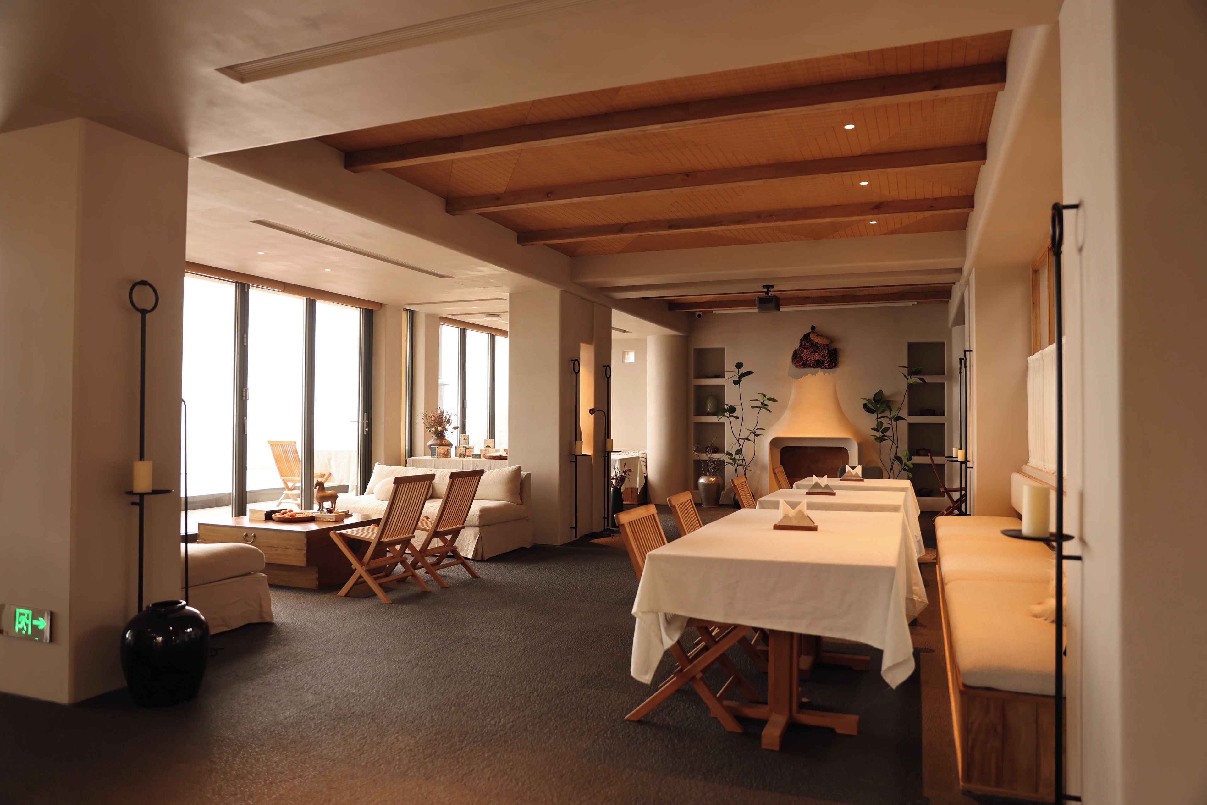 A spacious room with a high ceiling, large windows, and a dining area with a long table.