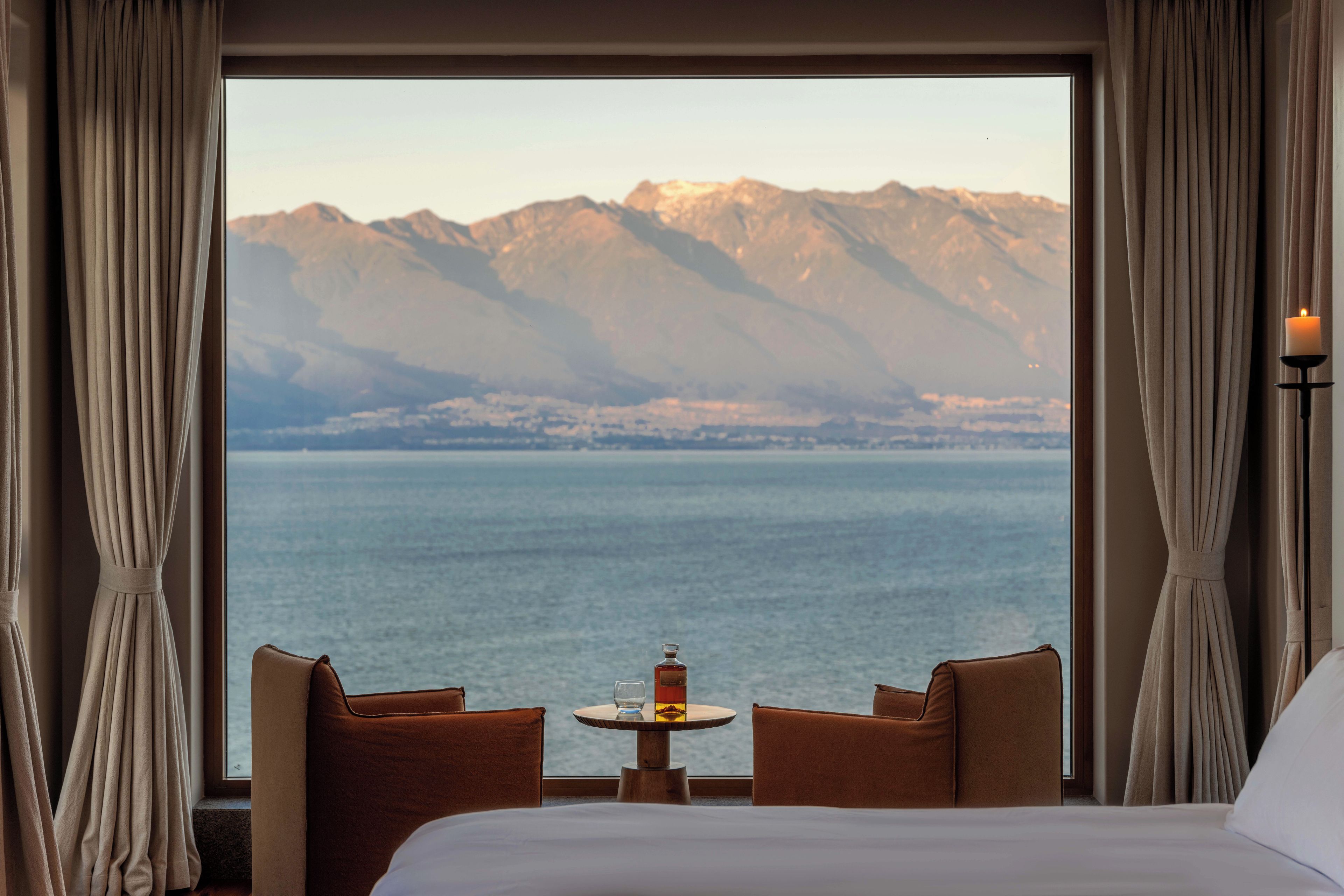A hotel room with a bed, two brown armchairs, a small round table with a candle, and a view of mountains and water.