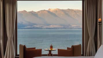 A hotel room with a bed, two brown armchairs, a small round table with a candle, and a view of mountains and water.
