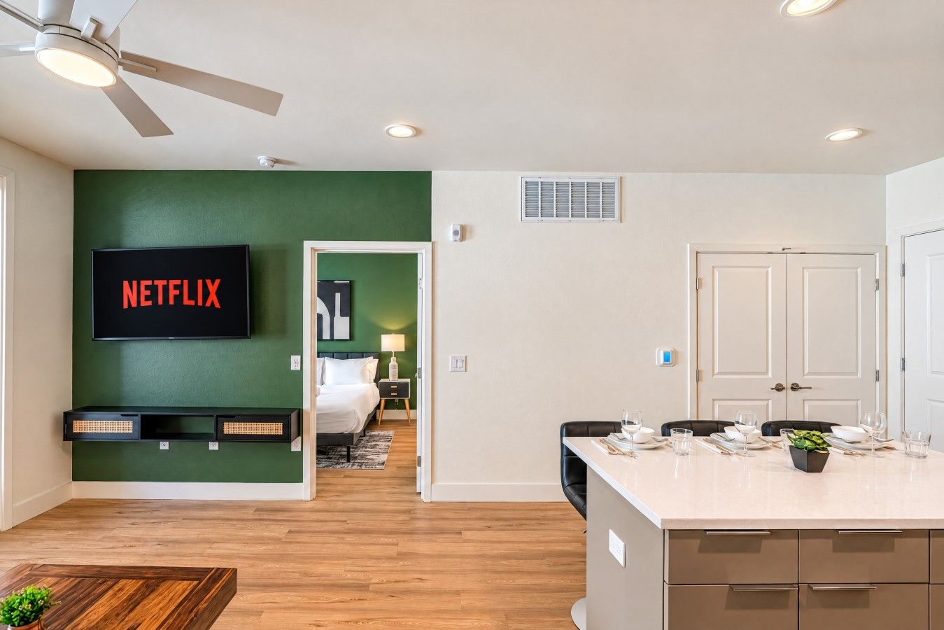 A modern hotel room with a green accent wall, a flat-screen TV, a bed, a desk, and a dining area.