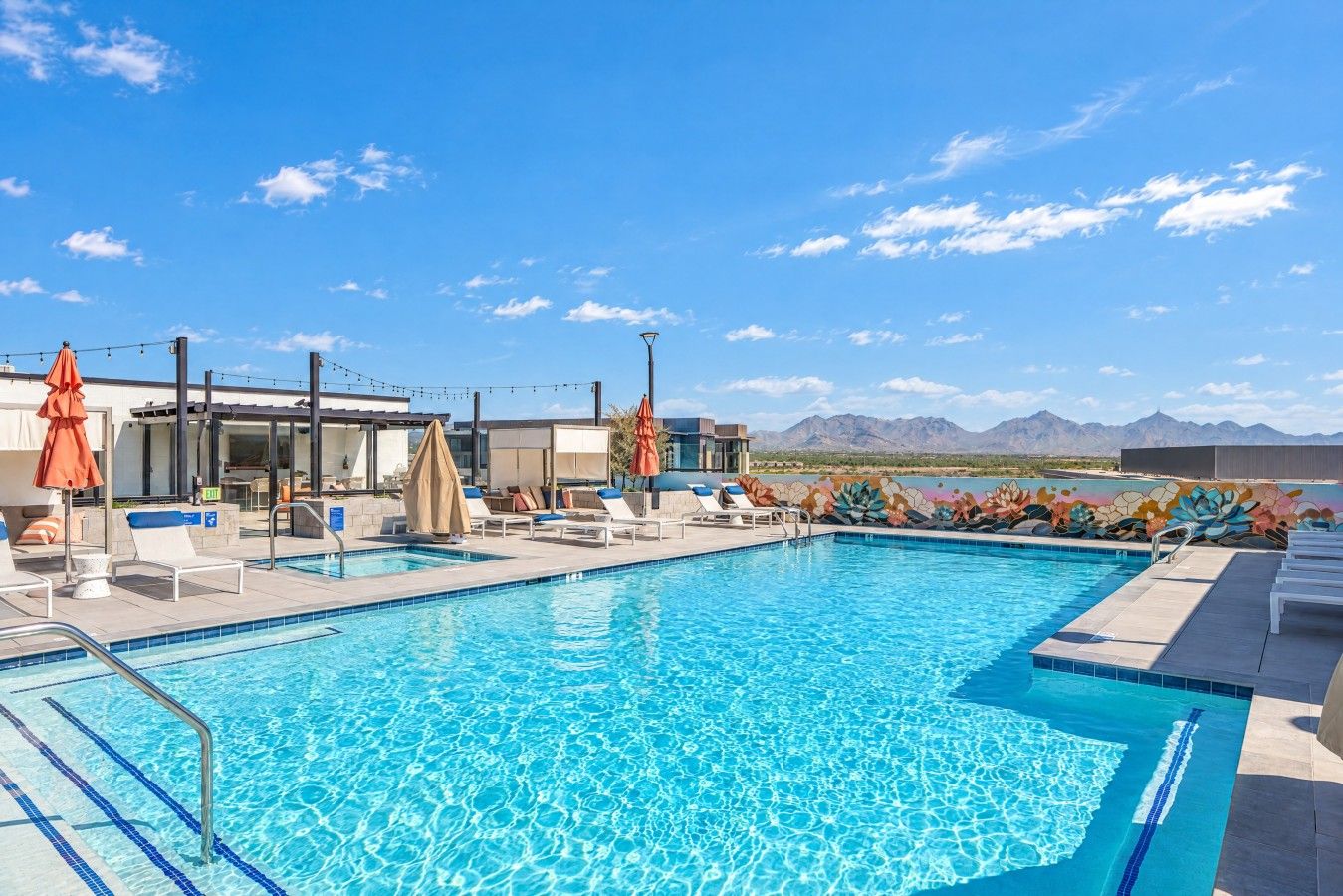 Un área de piscina con sillas de descanso y sombrillas, cielo despejado y montañas al fondo.