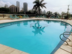 A swimming pool with a tiled edge, surrounded by lounge chairs and palm trees, with tall buildings in the background. - A12 en El Laguito: cerca del mar, piscina y aire acondicionado (CARTAGENA DE INDIAS)